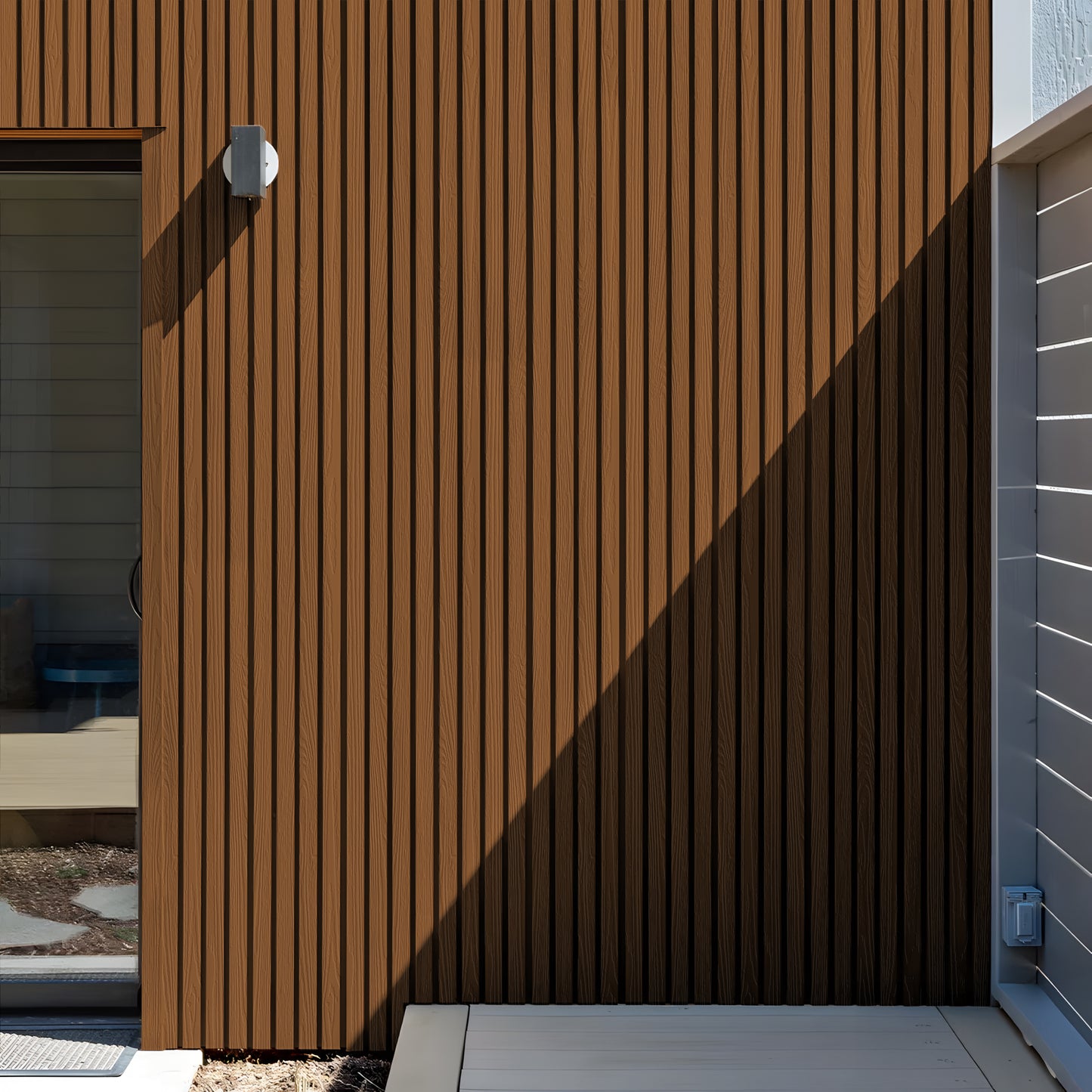 A Honey Chestnut Exterior WPC Panel by Landmark Decor forms a vertical slat wall with weather-resistant panels, paired with a wall light beside a glass door and white deck, partly shaded by sunlight.