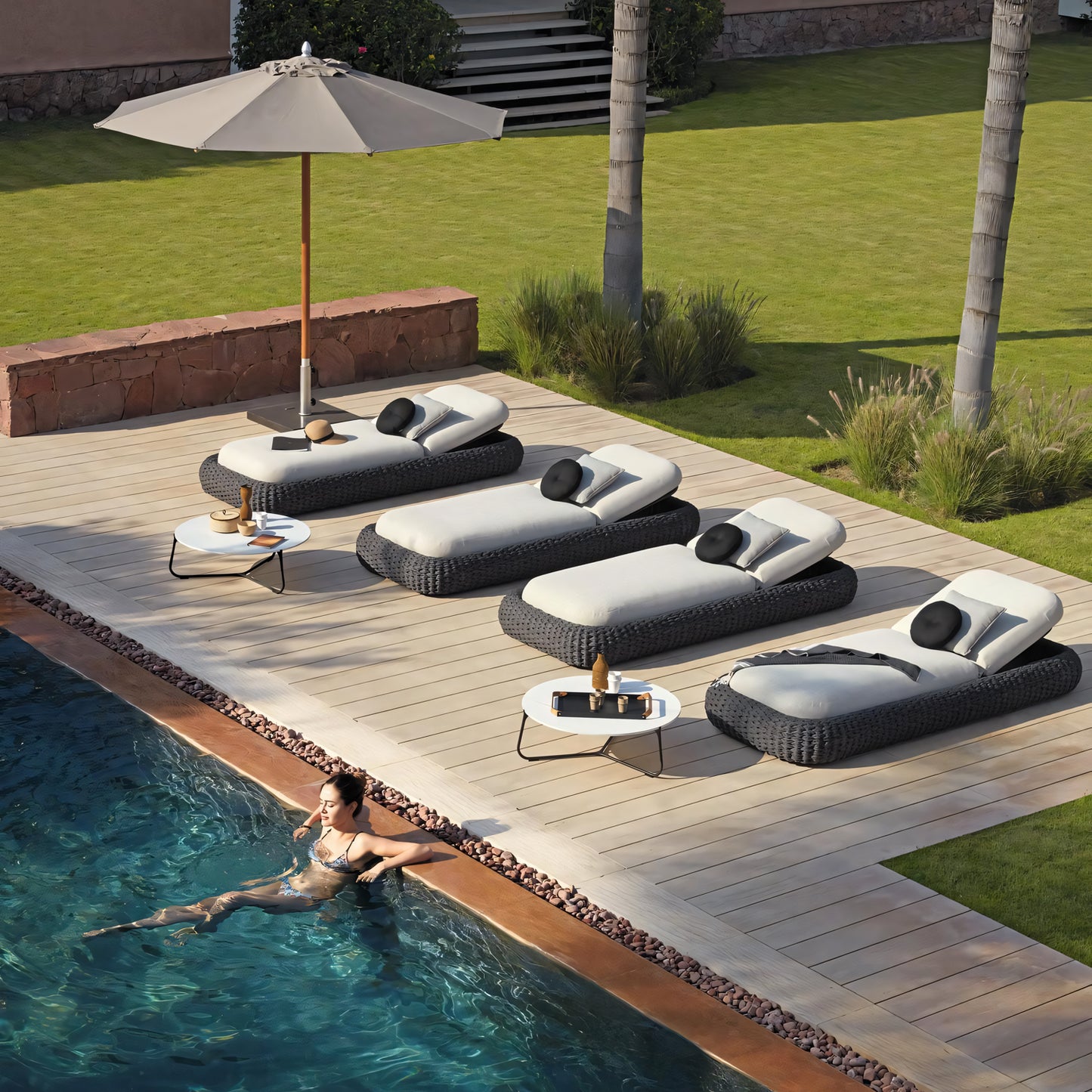 A woman in a swimsuit relaxes in a pool next to a wooden deck, enhancing her outdoor living experience. Nearby, four Palermo Terra Loungers by Landmark Decor, with beige cushions and black pillows, offer premium comfort. A large umbrella provides shade while drinks and a book rest on the small table.