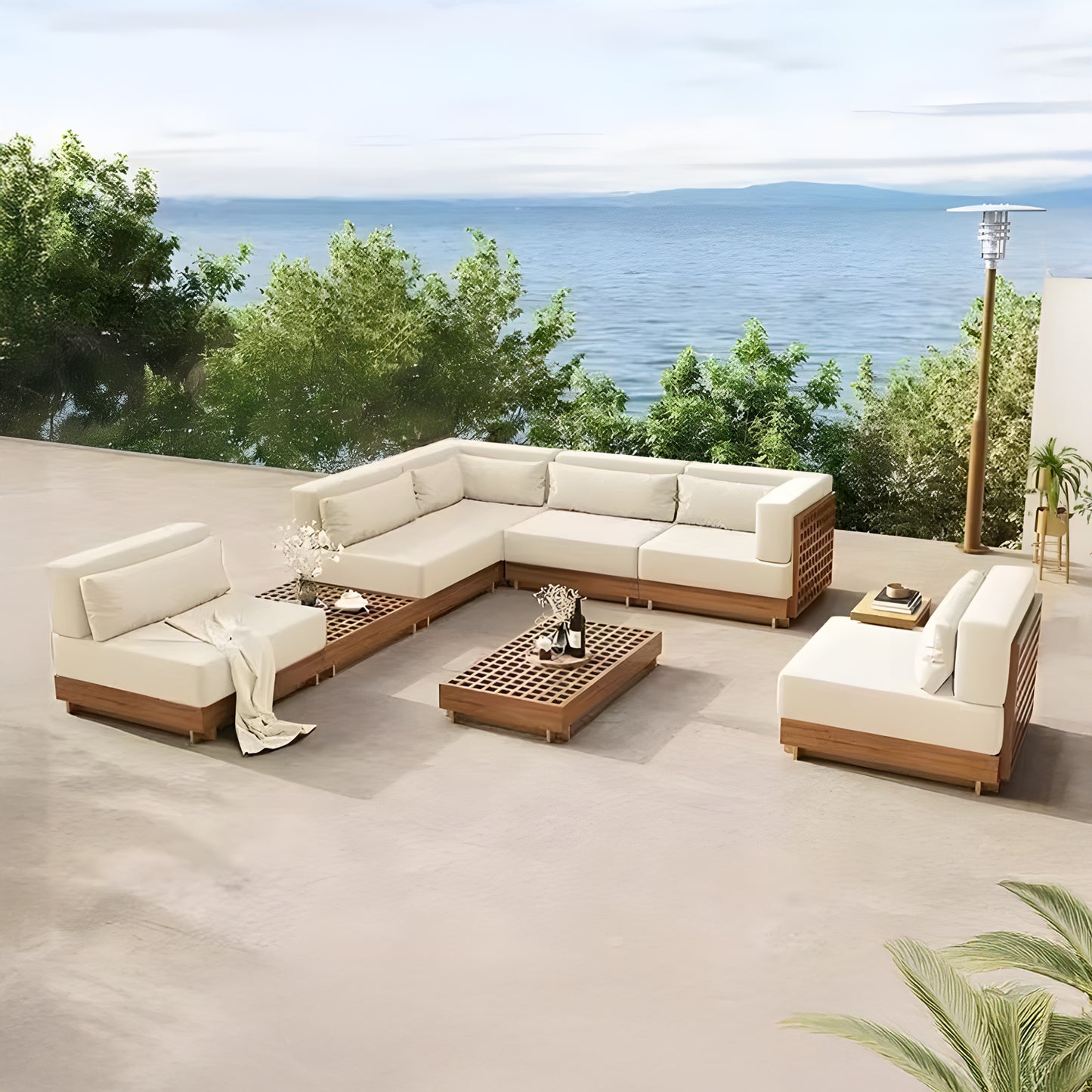 The Outdoor patio features the Santorini Serenity Collection by Landmark Decor, with beige all-weather frame sectional sofas and a matching coffee table arranged in a square. Set on a spacious terrace, it overlooks water amid lush greenery beneath blue skies.