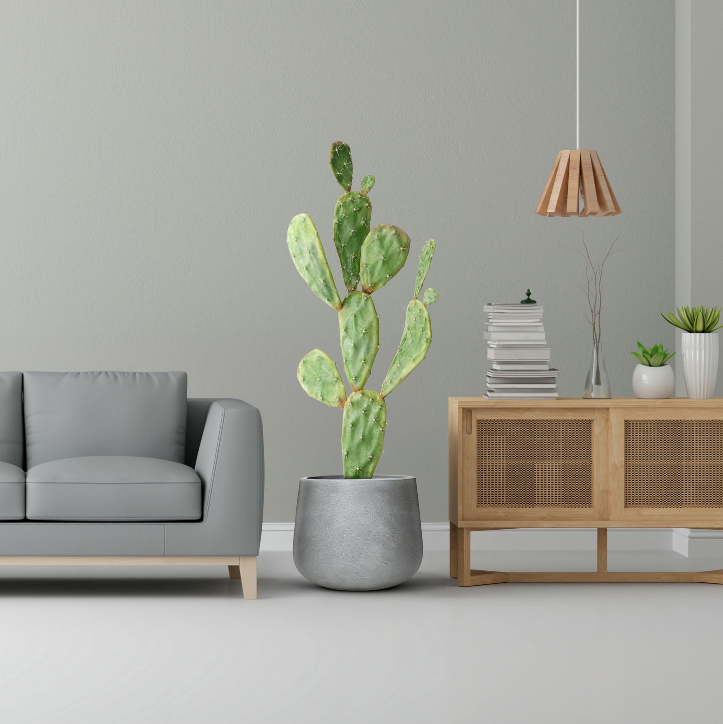 A modern living room boasts a light gray sofa, a large potted cactus in Landmark Decors Fortezza Planter - Small on the floor, and a wooden cabinet with books and plants. A pendant light enhances the soft green wall for added luxury and nature.