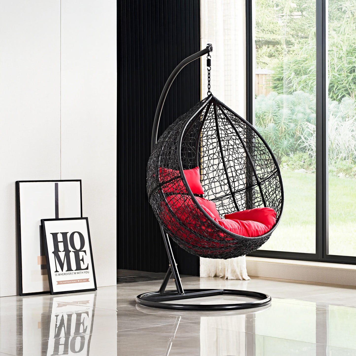 In a modern room, the Chelo Outdoor Swing Chair by Landmark Decor swings gently beside a large window, its red cushion adding luxury. A framed HOME text art leans against the wall, while the shiny floor mirrors the verdant outdoor view.