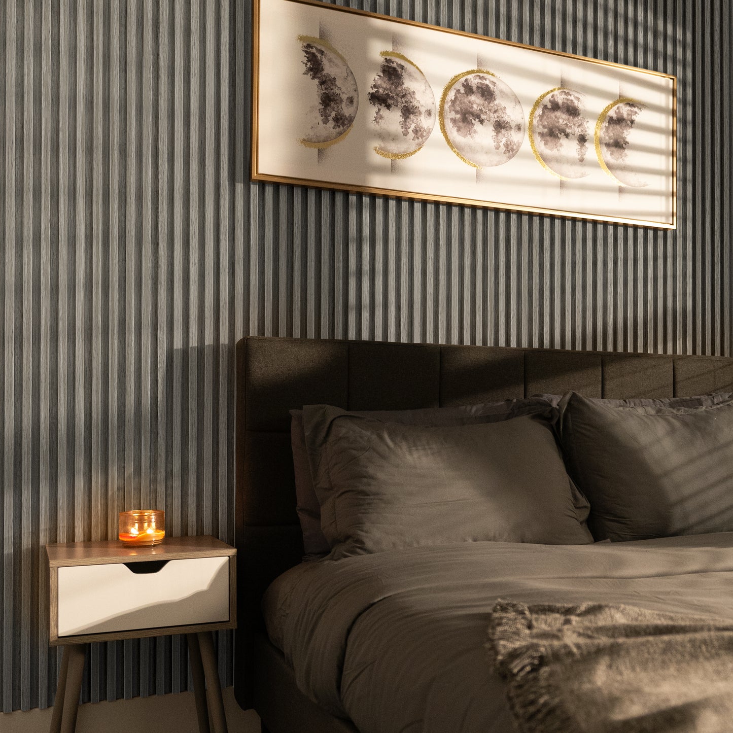 A neatly made bed with gray bedding sits beside a nightstand with a lit candle; framed moon phase artwork hangs above, complementing the Landmark Decor Ashwood Taupe WPC Fluted Slat Wall Panel.