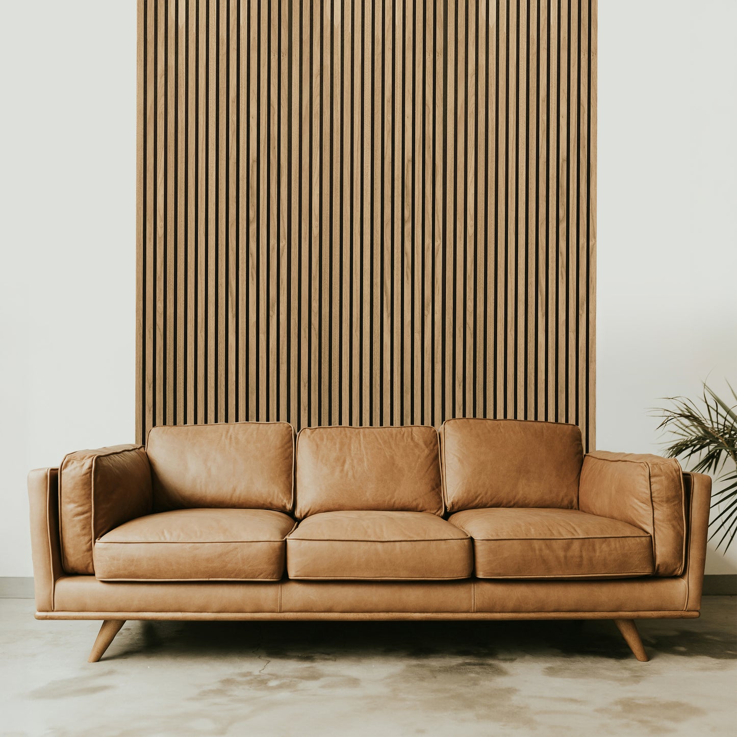 A tan leather sofa sits before a Landmark Decor Natural Oak Wood Color Acoustic Slat Wall Panel, with a small green plant on the right—a stylish choice for modern interiors.