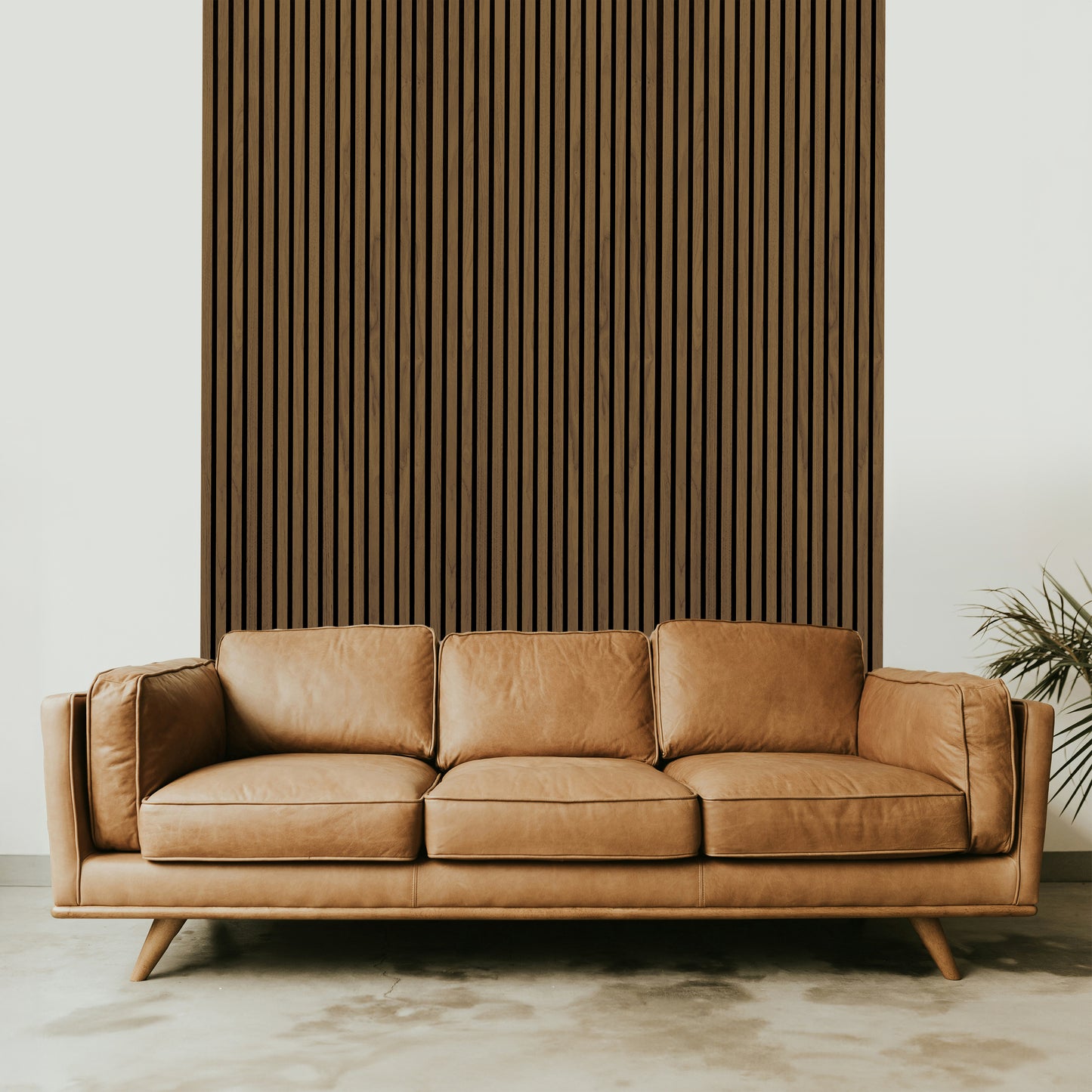 A tan leather three-seat sofa sits in front of a Landmark Decor Light Smoke Acoustic Slat Wall Panel, adding warmth and texture to the space, with a plant visible on the right edge.