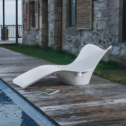 A Muse Astratta Lounger by Landmark Decor, featuring a sleek, curved modern white design, graces a wooden deck by the pool. A magazine lies nearby. The backdrop includes a stone wall with windows and closed shutters, crafting an exquisite outdoor retreat.