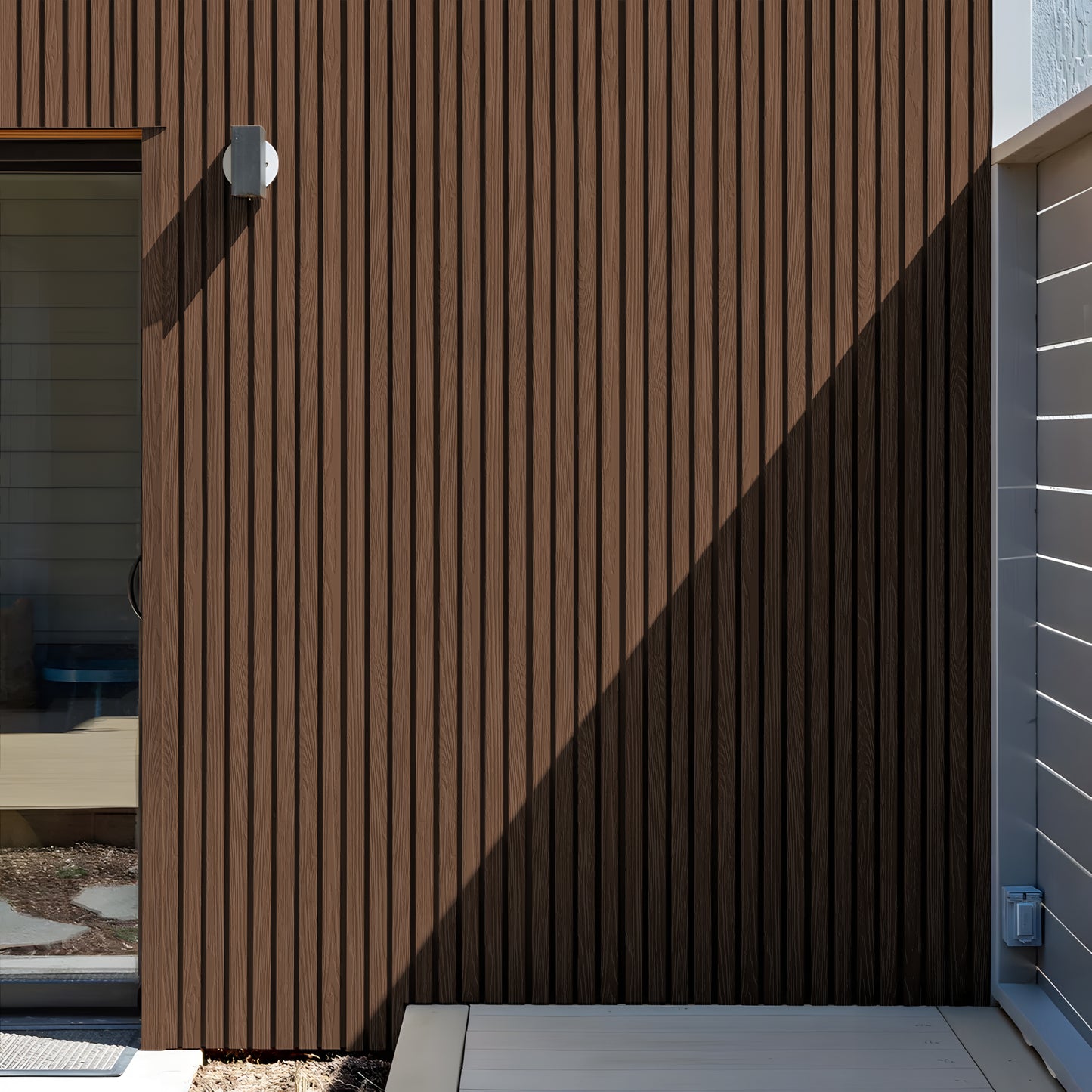 A Burnt Caramel Exterior WPC Panel by Landmark Decor features brown vertical slats, weather-resistant panels, a light fixture, partial glass door, and a distinct shadow sharply dividing sunlight and shade.