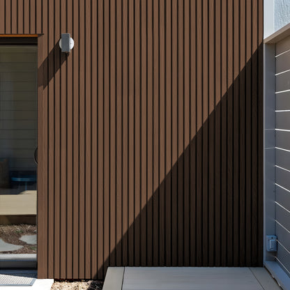 A Burnt Caramel Exterior WPC Panel by Landmark Decor features brown vertical slats, weather-resistant panels, a light fixture, partial glass door, and a distinct shadow sharply dividing sunlight and shade.