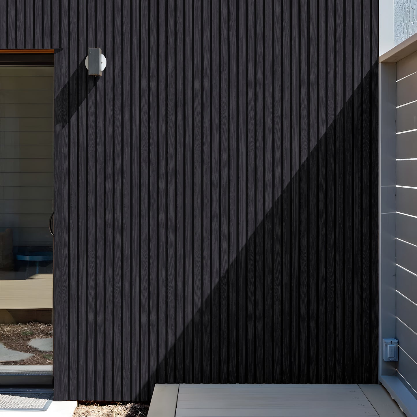 A modern exterior wall showcases Landmark Decor’s Charcoal Ash Exterior WPC Panels in vertical siding, a compact wall light, a glass door on the left, and shadows across the facade.