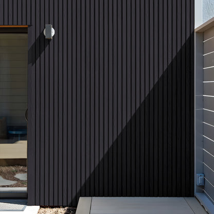 A modern exterior wall showcases Landmark Decor’s Charcoal Ash Exterior WPC Panels in vertical siding, a compact wall light, a glass door on the left, and shadows across the facade.