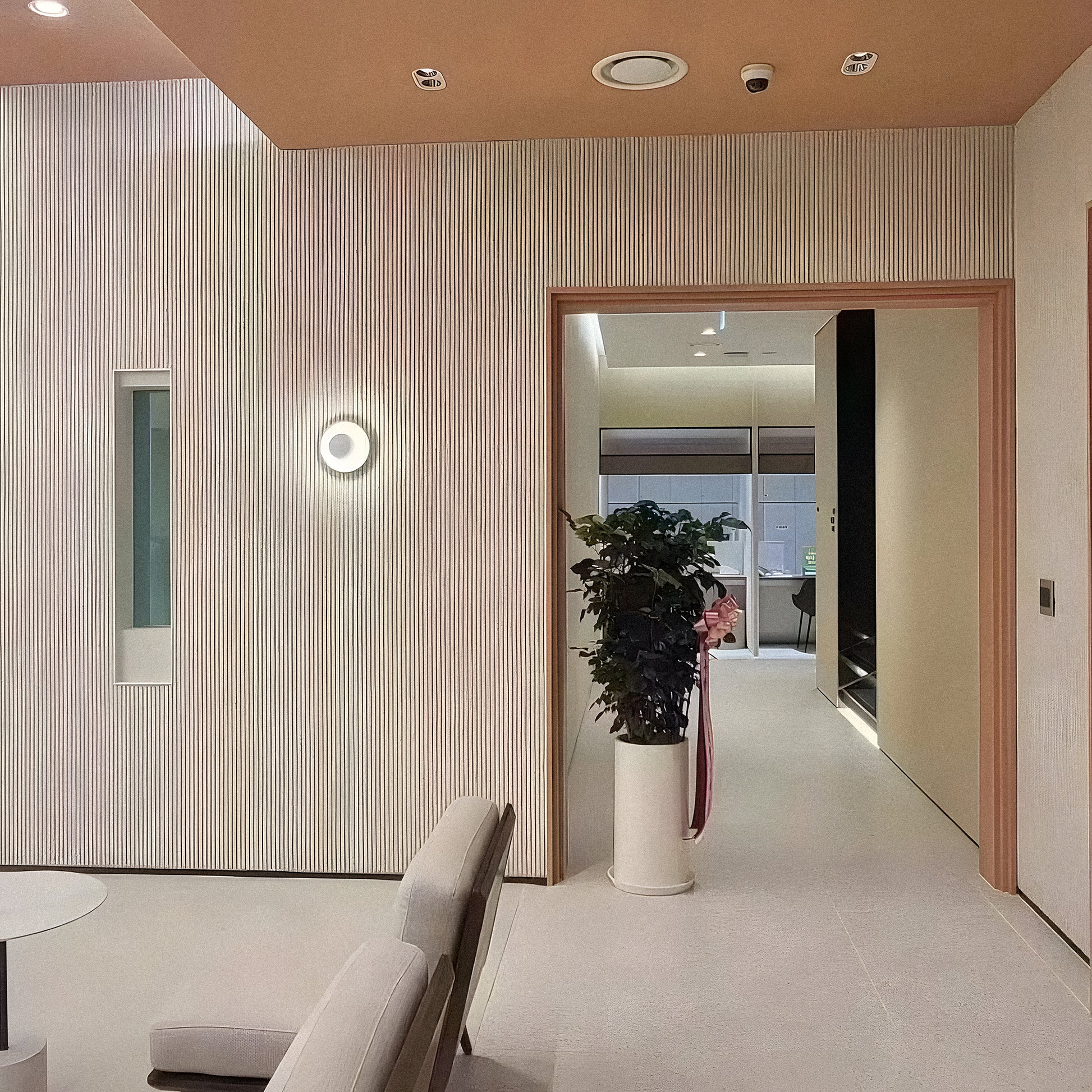A modern minimalist interior features beige walls, a round light fixture, a tall potted plant, and a doorway. Lightweight Oman Linear Stone Sheet - Grey by Landmark Decor accents the space, with two light chairs partially visible in front.
