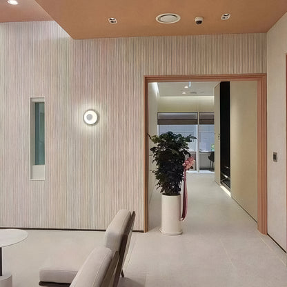 A modern minimalist interior features beige walls, a round light fixture, a tall potted plant, and a doorway. Lightweight Oman Linear Stone Sheet - Grey by Landmark Decor accents the space, with two light chairs partially visible in front.