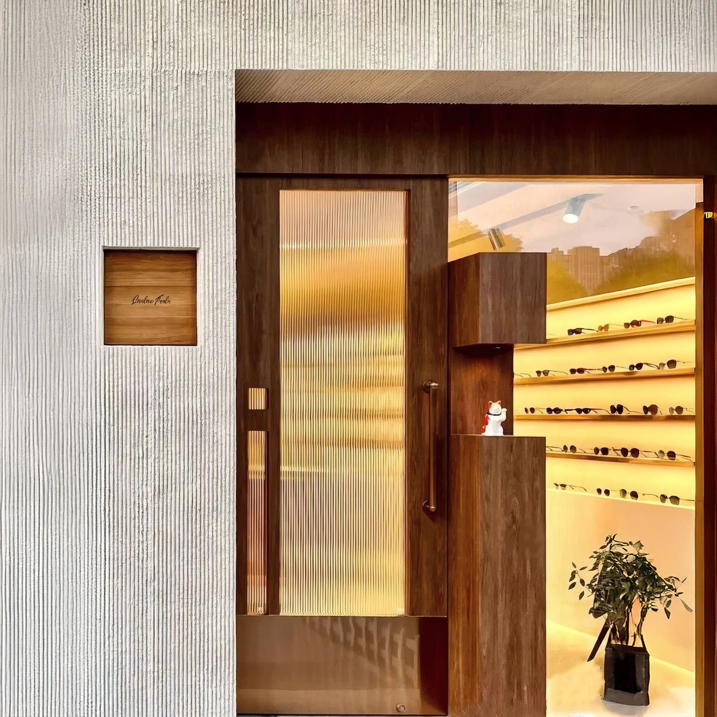 Modern storefront with a textured white exterior featuring Landmark Decor's Oman Linear Stone Sheet - Grey, a wooden door, glass panels, a small potted plant, and illuminated shelves displaying eyewear inside.