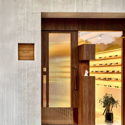 Modern storefront with a textured white exterior featuring Landmark Decor's Oman Linear Stone Sheet - Grey, a wooden door, glass panels, a small potted plant, and illuminated shelves displaying eyewear inside.