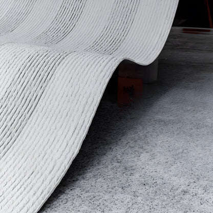 The Oman Linear Stone Sheet - Grey by Landmark Decor features a curved, ribbed flexible stone veneer arching above a textured gray floor, creating an industrial look.