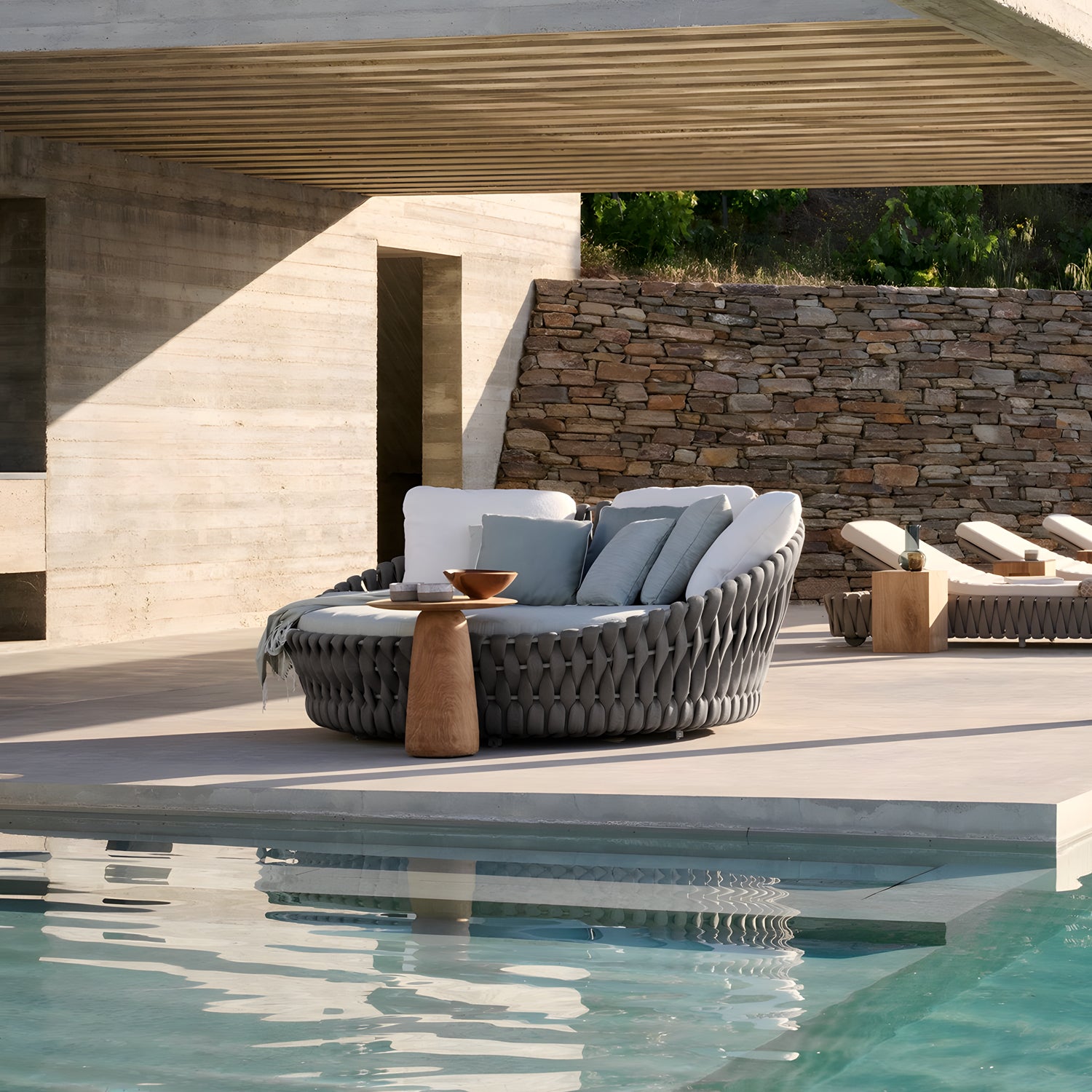 Experience a luxurious retreat with Landmark Decors Mediterranea Luxe Lounge by the pool. The setting includes a round woven sofa with cushions, a small wooden table with a decorative bowl, stone walls, and chaise lounges for a modern, serene outdoor oasis.