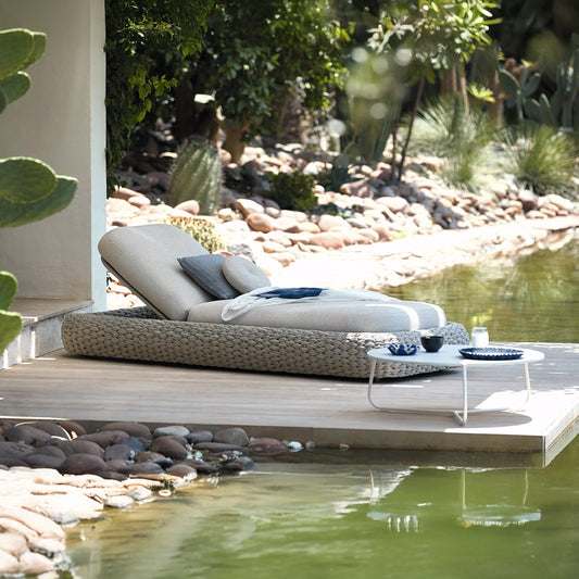 A Marbella Retreat Lounger by Landmark Decor sits on a wooden deck by the pond, surrounded by rocks and greenery. A small white table holds a bowl and glass. Cacti and various plants complete this luxurious retreat for ideal outdoor living.