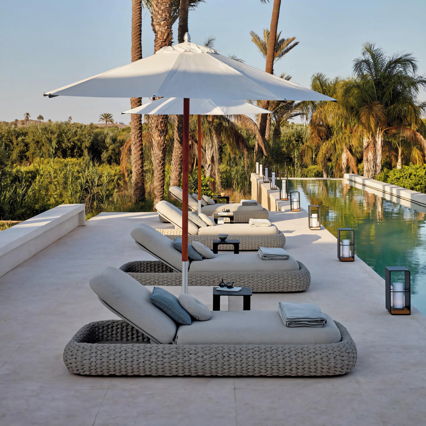The Marbella Retreat Lounger by Landmark Decor offers a luxurious escape with modern chaise lounges and white umbrellas lined beside a sleek pool, complete with small tables and lanterns. Surrounded by palm trees and lush greenery under a clear sky, it truly captures the essence of outdoor living.