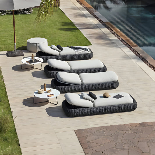 The outdoor area showcases four Landmark Decor Palermo Terra Loungers crafted from woven rattan, featuring white cushions and black headrests on a light wooden deck. Two round tables with decor sit near the pool, surrounded by lush grass and vibrant plants.