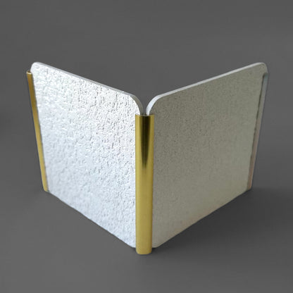 The Landmark Decor PVC Wall Inner Corner Strip features two textured silver panels joined at a right angle by gold-colored cylindrical hinges with precise edges, displayed on a gray surface.