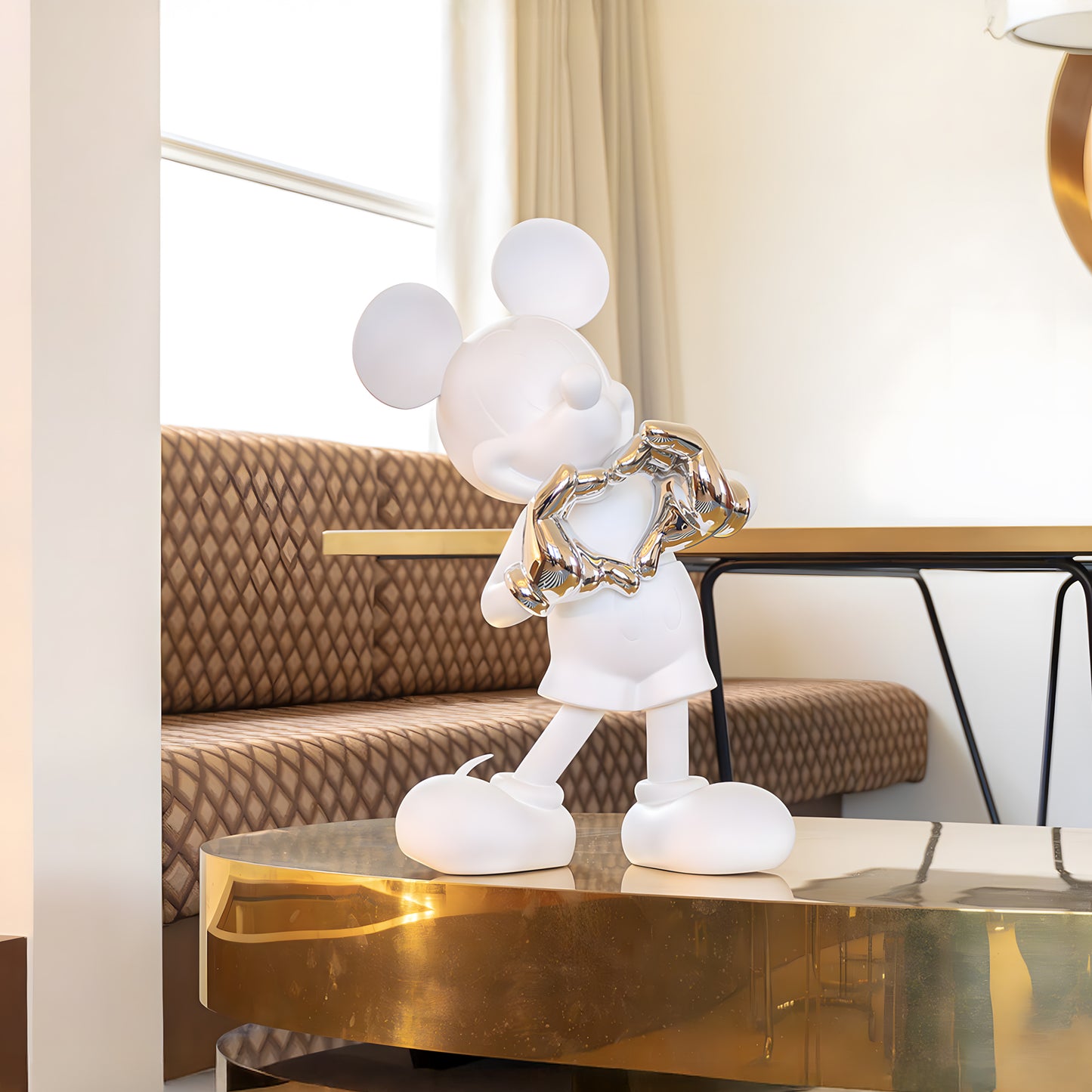 The Heart U - Small by Landmark Decor is a white Mickey Mouse sculpture with metallic hands forming a heart, featured as a striking accent on a gold table in a contemporary room with patterned seating and neutral decor.