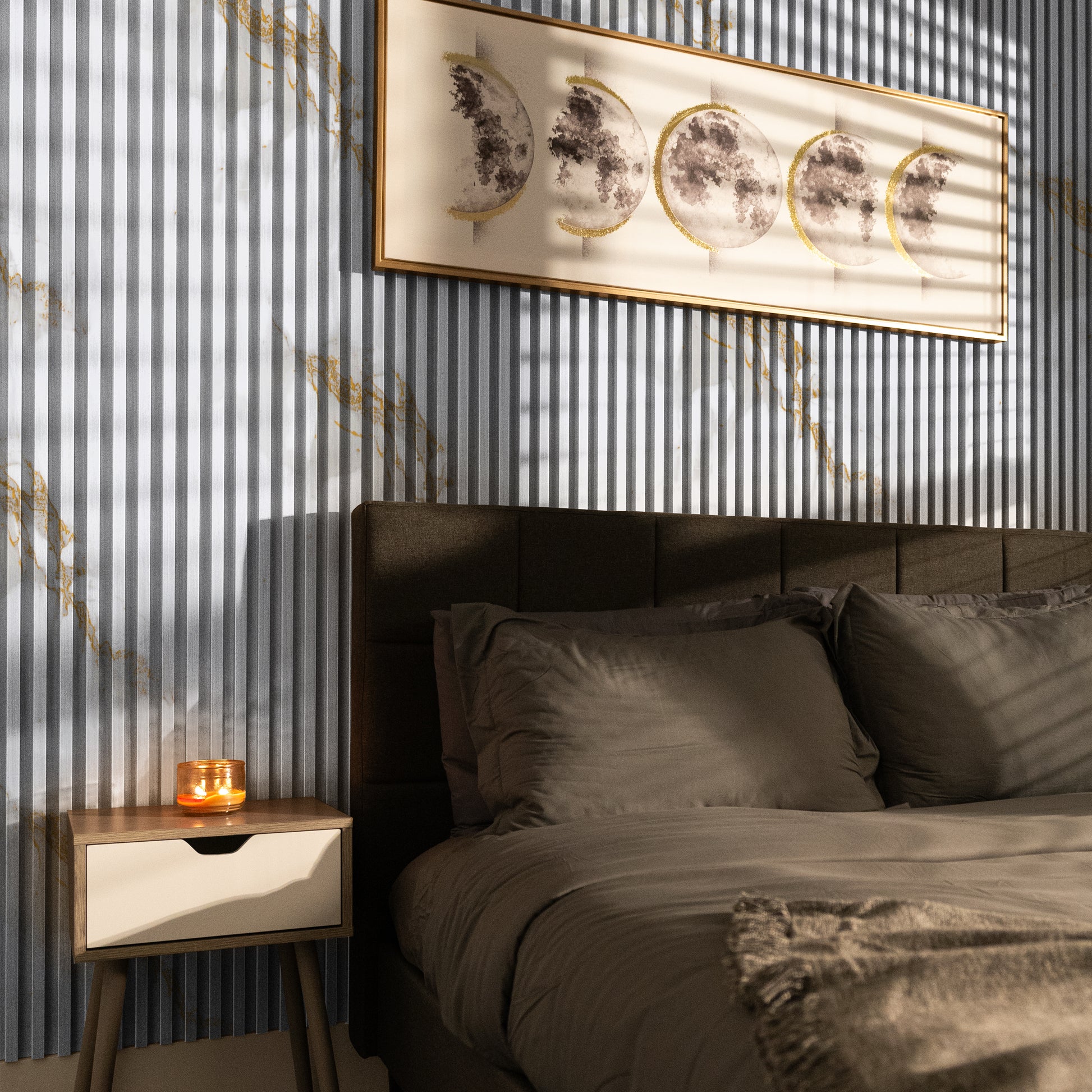 A modern bedroom with a gray bed, brown bedding, and a wooden nightstand with a lit candle, features framed lunar phase art on accent walls clad in Landmark Decor’s Calacatta Dawn WPC Fluted Slat Wall Panel as sunlight creates gentle shadows.