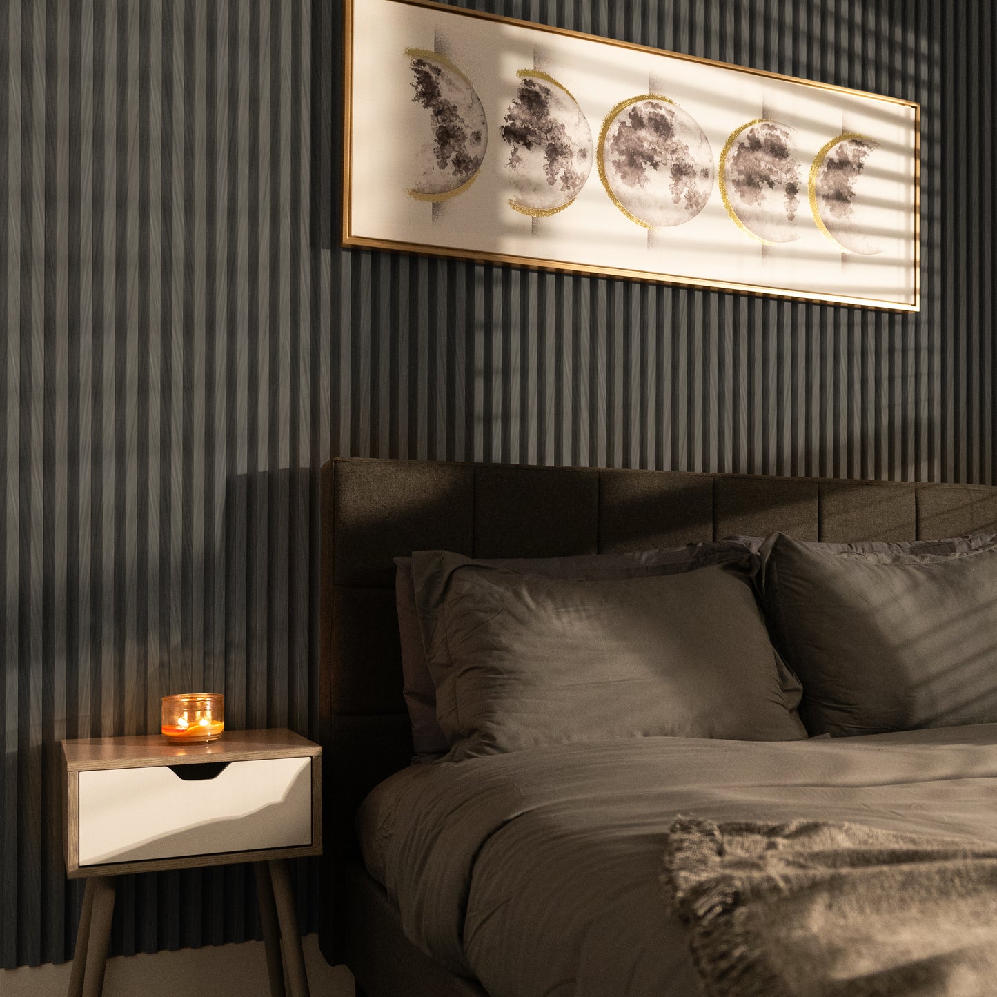 A neatly made bed with gray bedding, a wooden nightstand with a lit candle, and moon phase artwork highlight the Ebony Chevron WPC Fluted Slat Wall Panel by Landmark Decor on the dark textured accent wall.