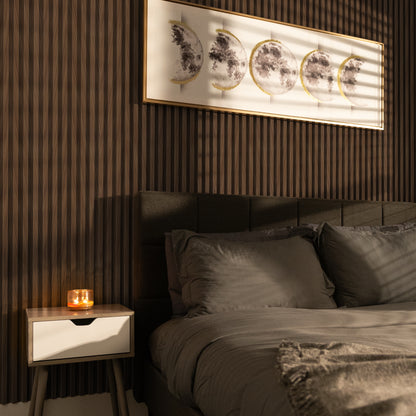 A modern bedroom featuring Landmark Decor's Cognac Chevron WPC Fluted Slat Wall Panel accent wall, a gray upholstered bed with matching bedding, a candle on a small nightstand, and framed moon phase art above the bed.