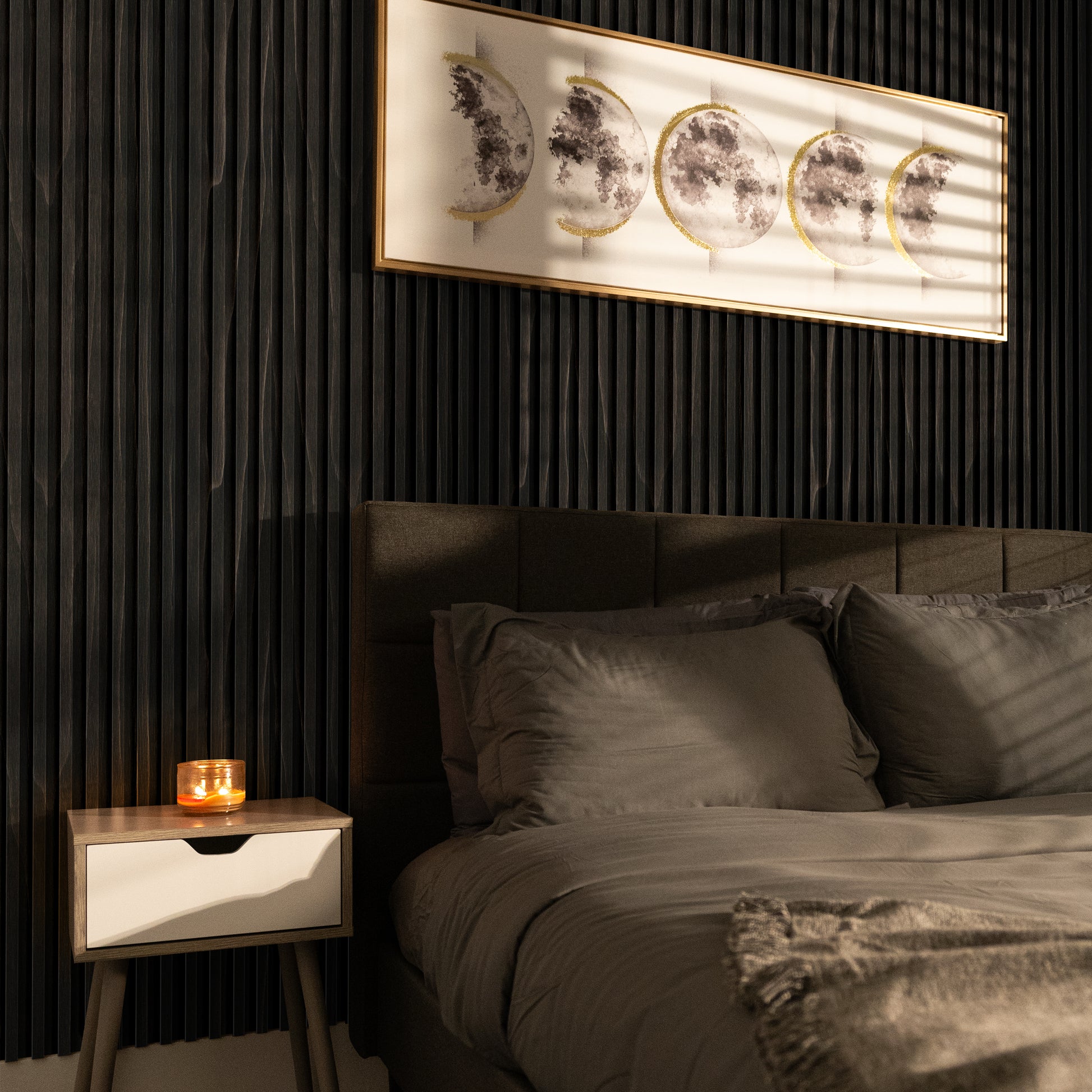 A neatly made bed with gray linens sits by a nightstand holding a lit candle, set against a dark accent wall featuring framed moon phase art and Landmark Decor's Smoked Walnut Flow WPC Fluted Slat Wall Panel for added depth.