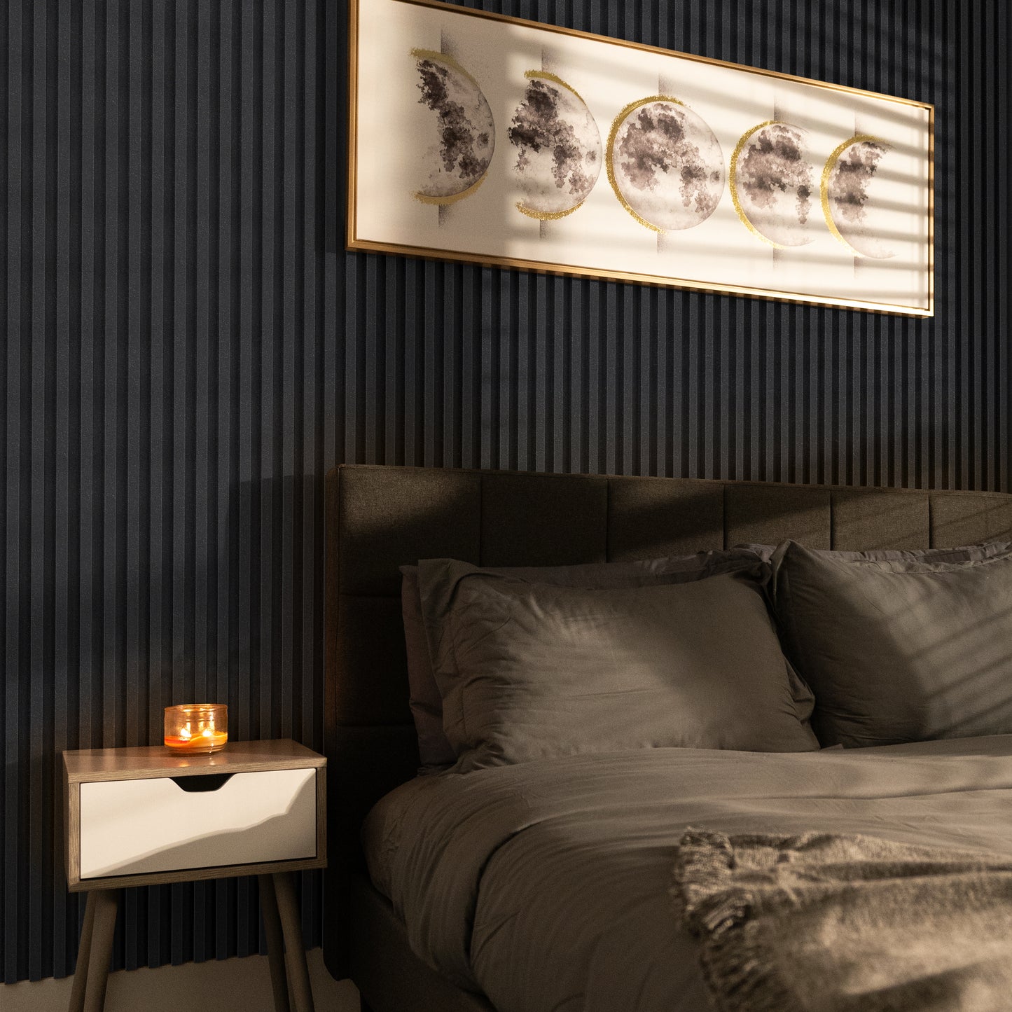 A modern bedroom features a grey bed, nightstand with a lit candle, and framed moon phase art, all set against a Landmark Decor Obsidian Shadow WPC Fluted Slat Wall Panel accent wall.