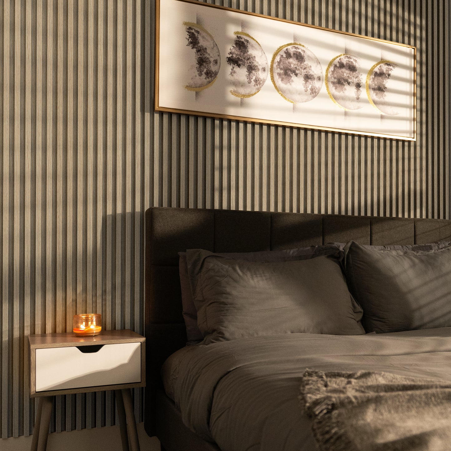 A neatly made bed with gray bedding sits beside a nightstand and lit candle, beneath framed moon phase art on a striped accent wall featuring Landmark Decor’s Bamboo Grain WPC Fluted Slat Wall Panel.