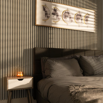A neatly made bed with gray bedding sits beside a nightstand and lit candle, beneath framed moon phase art on a striped accent wall featuring Landmark Decor’s Bamboo Grain WPC Fluted Slat Wall Panel.