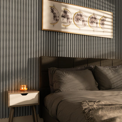 A neatly made bed with gray bedding sits beside a nightstand with a lit candle; framed moon phase artwork hangs above, complementing the Landmark Decor Ashwood Taupe WPC Fluted Slat Wall Panel.