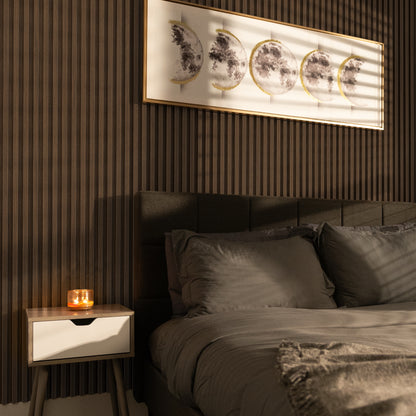 A neatly made bed with gray bedding stands beside a candle-lit nightstand; moon phase art hangs on Landmark Decor’s Toasted Walnut WPC Fluted Slat Wall Panel, giving character to the cozy space.