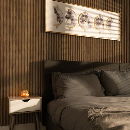 Modern bedroom featuring Landmark Decor's Golden Teakwood WPC Fluted Slat Wall Panel, moon phase artwork, a gray bed, and a lit candle on a nightstand. Sunlight creates striking shadows across the space.