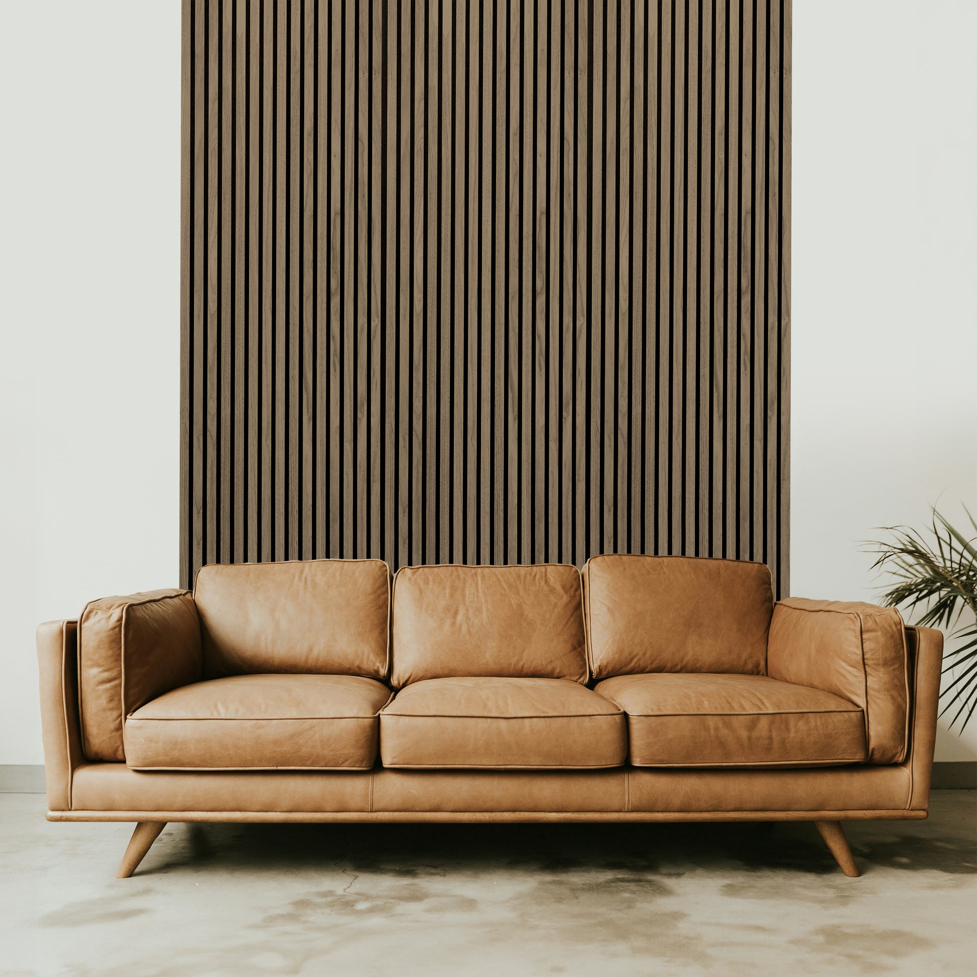 A tan leather sofa sits before a Landmark Decor Light Walnut Acoustic Slat Wall Panel, with a green plant partially visible on the right, adding style and warmth to the interior design.