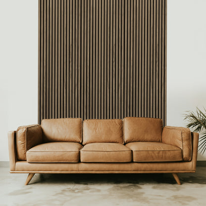 A tan leather sofa sits before a Landmark Decor Light Walnut Acoustic Slat Wall Panel, with a green plant partially visible on the right, adding style and warmth to the interior design.
