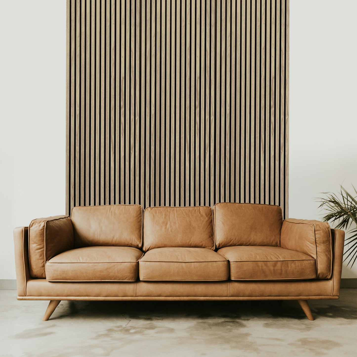 A tan leather sofa sits in front of Landmark Decor’s Light Oak Acoustic Slat Wall Panel, with a plant partially visible on the right—a stylish accent for modern interiors.