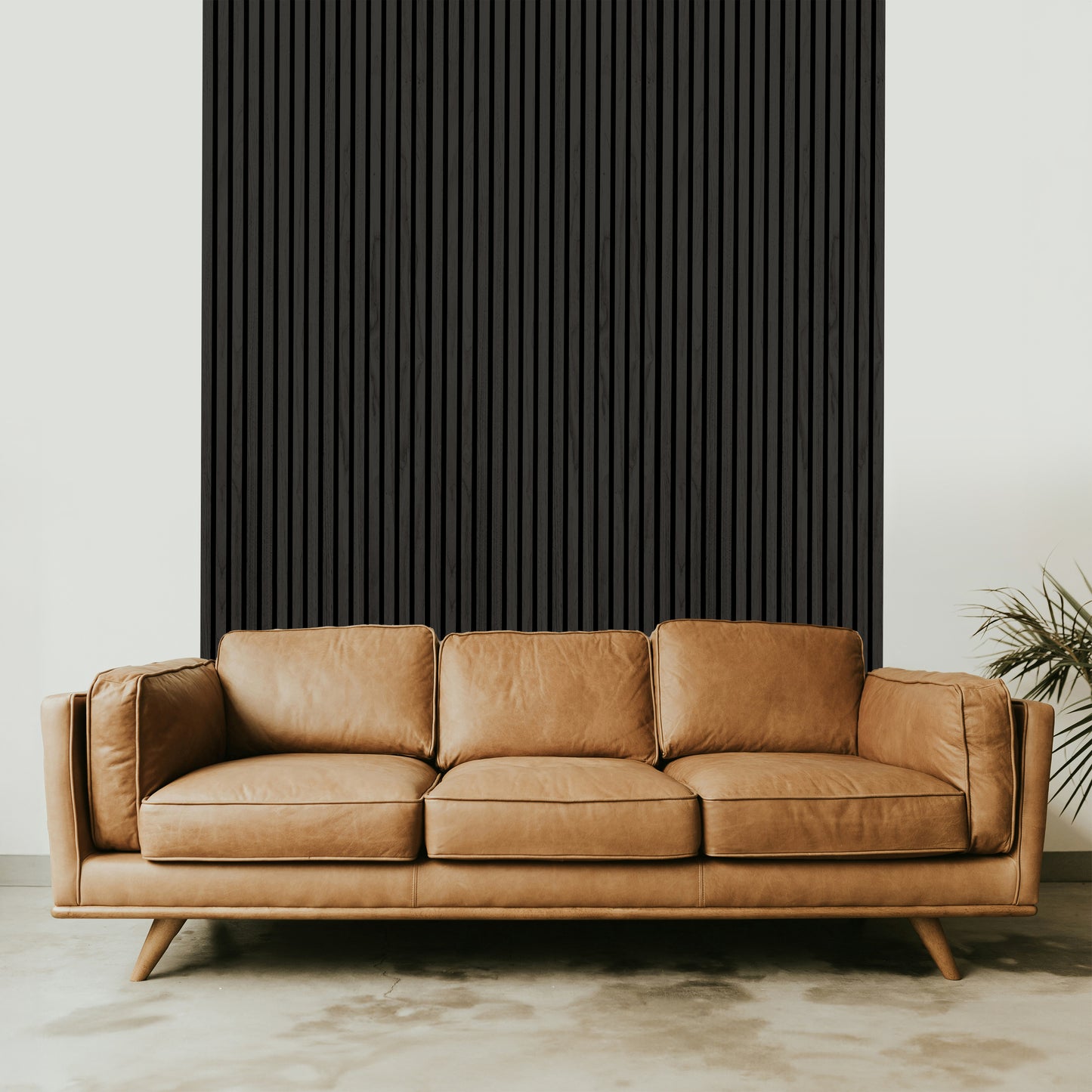 A tan leather sofa with three cushions sits in front of a Landmark Decor Black Ebony Acoustic Slat Wall Panel; a plant is partially visible on the right, enhancing the interior design.