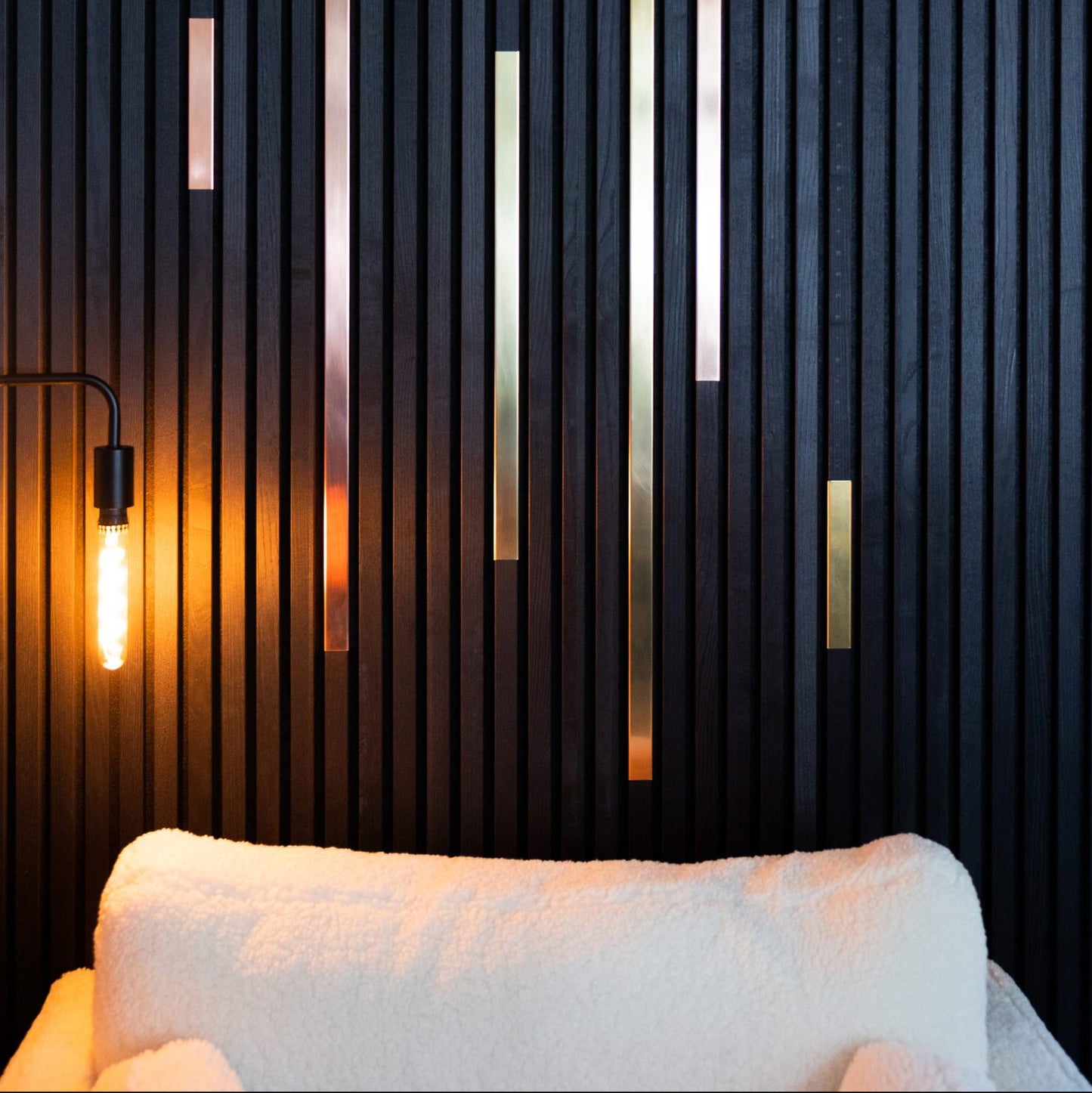 Experience seamless design flexibility with Landmark Decor's Wood Panel Metal Cover-Black, featuring modern vertical black wood panels with metallic gold accents, paired with a glowing bulb lamp and a plush white chair in the foreground.