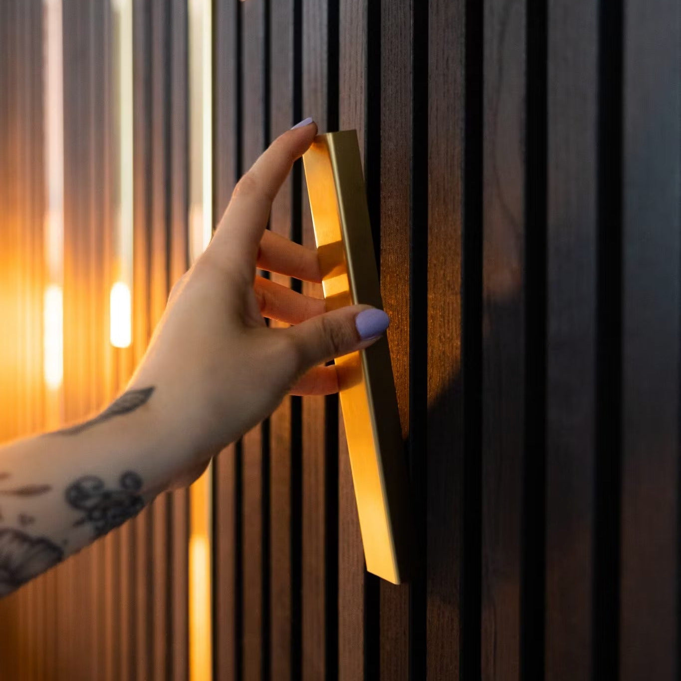 A hand with a floral tattoo and lavender nails grips a gold handle on Landmark Decor’s Wood Panel Metal Cover-Black, mounted on dark wood panels and enhanced by warm lighting for versatile design.