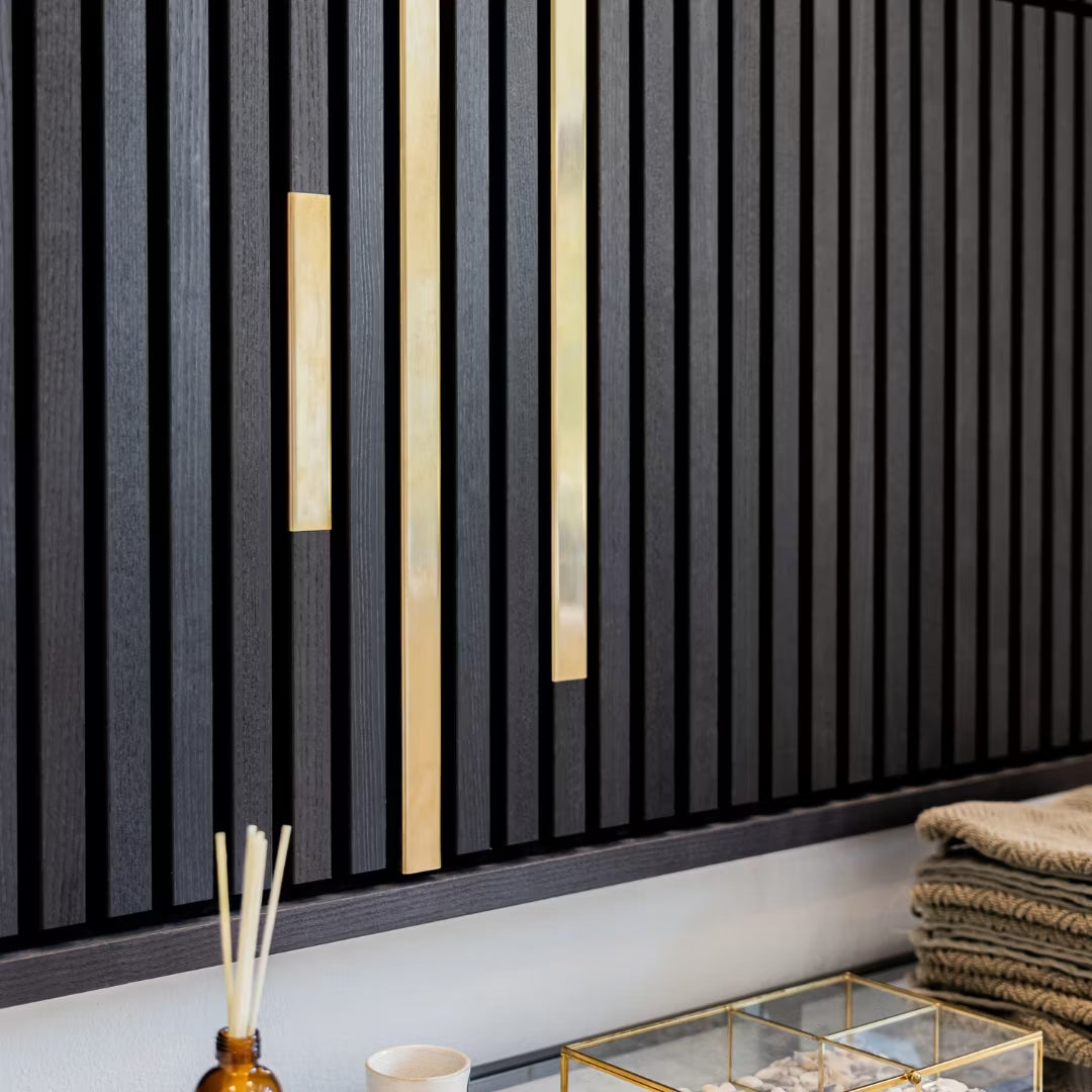 Landmark Decor’s Wood Panel Metal Cover-Black features black slatted panels with vertical gold accents, adding a modern touch above a counter displaying folded towels, a glass container, and a brown reed diffuser.
