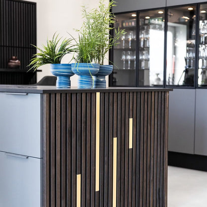 A modern kitchen island features Landmark Decor’s Wood Panel Metal Cover-Black with gold accents, topped with three potted green plants in blue striped pots. Glassware in cabinets adds to the space’s versatile design.